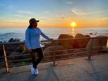 Rear view of man standing by railing against sea during sunset