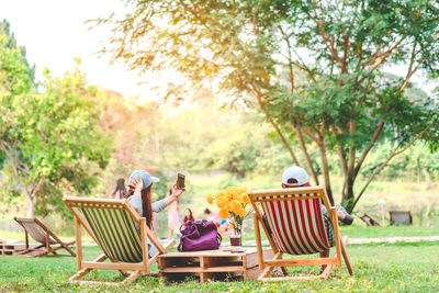 Rear view of woman taking selfie while sitting with man on deck chair at park