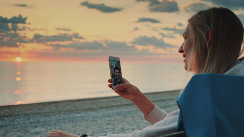 Woman using mobile phone at beach against sky during sunset