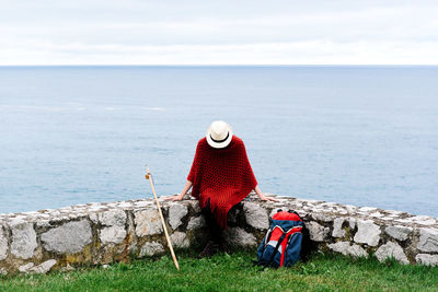 Happy young female backpacker in stylish clothes sitting on stone border against sea bay and looking down while resting during camino de santiago route in spain