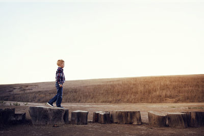 Side view of boy standing on wood at field against clear sky