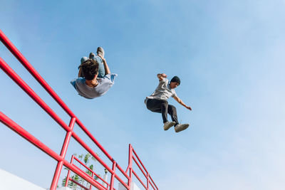 From below fearless male friends jumping above metal railing in city while performing parkour stunt on sunny day
