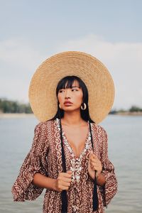 Young woman wearing hat looking away standing at beach against sky