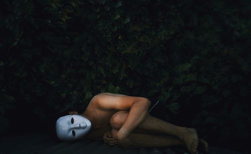 Shirtless man wearing white mask while lying on roof against plants