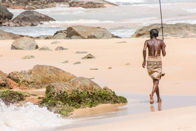 Rear view of shirtless fisherman running at beach towards sea on sunny day