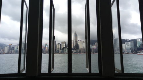 Modern buildings by river against cloudy sky seen through window