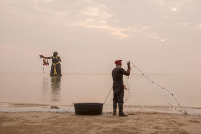 Men fishing on beach against sky during sunset