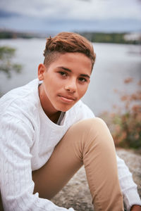 Portrait of confident teenage boy sitting on lakeshore in park during picnic