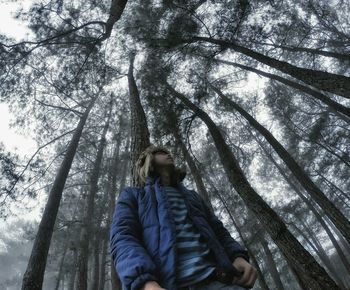 Low angle view of young man standing amidst trees at forest during foggy weather