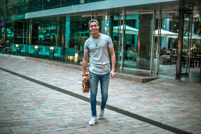 Portrait of young man carrying guitar on footpath against building