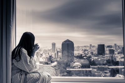 Woman drinking coffee while looking at cityscape through window