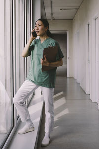 Full length of female physician holding clipboard while talking on smart phone in hospital corridor