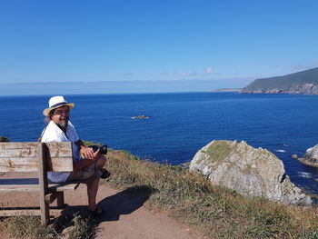 Portrait of man sitting on bench at mountain by sea against sky