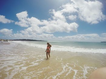 Woman wearing bikini walking on shore at beach against cloudy sky