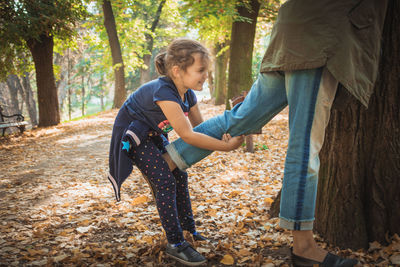 Daughter holding leg of mother standing by tree trunk in park