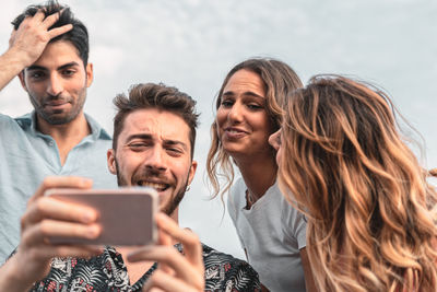 Close-up of cheerful young man showing smart phone to friends against sky