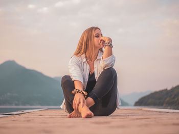 Young woman sitting on shore against sky during sunset