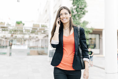 Smiling young talking on mobile phone while walking on footpath at city