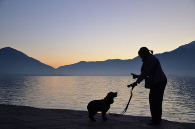 Silhouette woman with dog standing by lake against mountains and clear sky at dusk