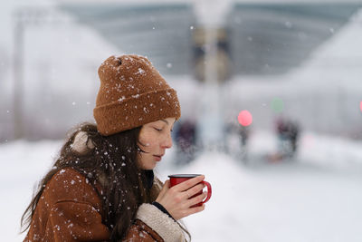 Asian girl traveler drinking hot tea trying to keep warm on city railway station on snowy winter day