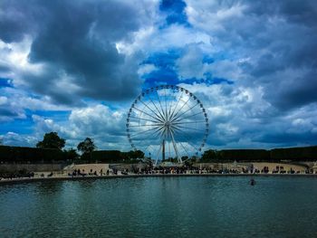 Ferris wheel by river against cloudy sky