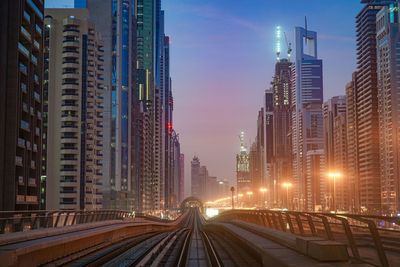 Illuminated railroad tracks amidst buildings in city against sky