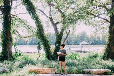 Full length of young woman sitting on tree trunk by lake against sky
