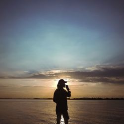 Silhouette woman photographing on beach against sky during sunset