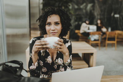 Portrait of woman having coffee while sitting by laptop computer at table in cafe