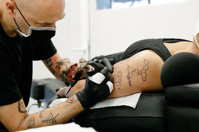 Crop male tattooist with machine making tattoo on arm of woman lying on table with closed eyes in salon