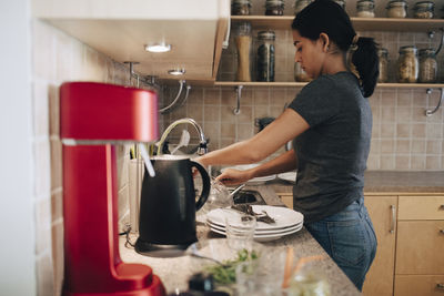 Side view of woman washing utensils in kitchen sink at home