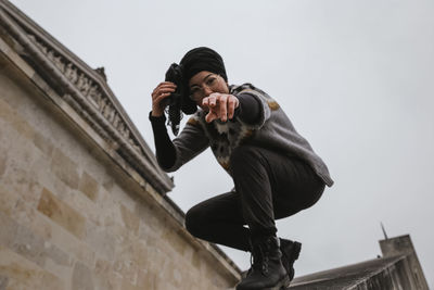 Low angle portrait of woman gesturing while crouching in city against sky