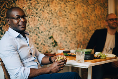 Portrait of man having food while sitting with friend by wall at home