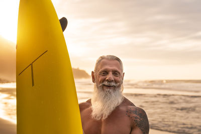 Portrait of shirtless senior man holding surfboard at beach against sky