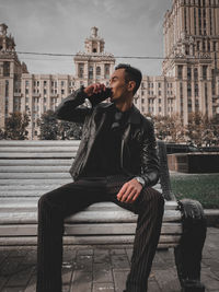 Young man looking away while sitting on seat in city