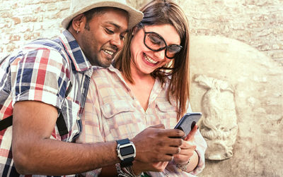 Cheerful couple using phone while standing against wall