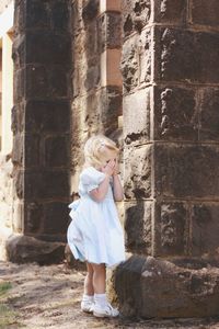 Full length of cute girl covering face while standing by wall