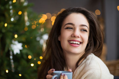 Smiling young woman holding coffee cup looking away against christmas tree