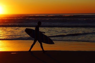 Silhouette man with surfboard walking on shore at beach against sky during sunset