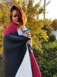 Thoughtful young woman wrapped in blanket while having drink against plants