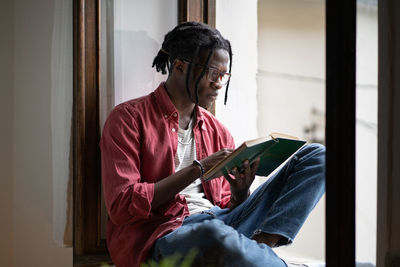 Focused african student guy nerd in glasses sitting on windowsill reading book, preparing for exam