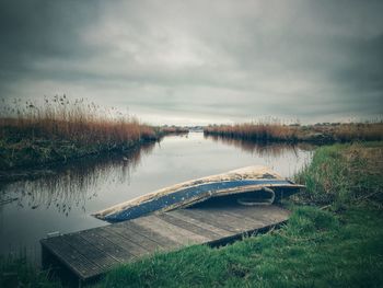 Old boat at jetty by river against cloudy sky