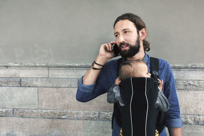 Mid adult man using mobile phone while carrying son in baby carrier against wall
