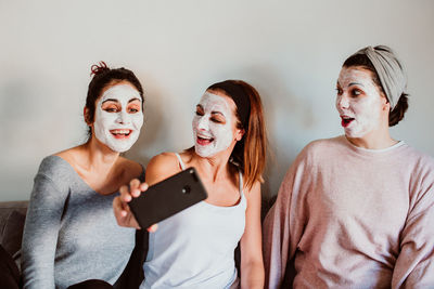 Female friends with facial masks taking selfie with against wall at home