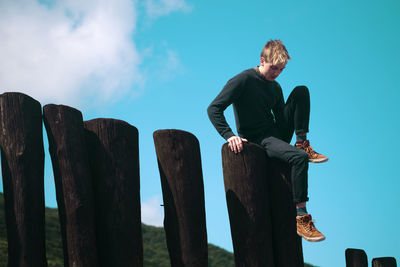 Low angle view of young man sitting on wood against sky