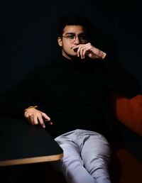 Portrait of confident young man in eyeglasses sitting in dark 