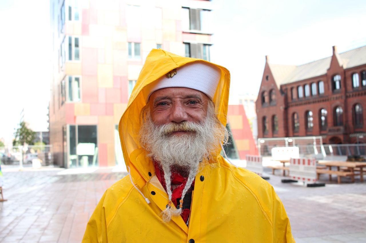 portrait, building exterior, looking at camera, front view, architecture, built structure, one animal, city, dog, incidental people, animal themes, street, focus on foreground, outdoors, yellow, close-up, day, beard