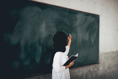 Teacher holding chalk while standing against blackboard in classroom