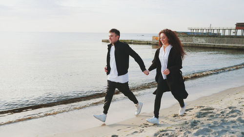 Full length of young couple holding hands while walking at beach against sky