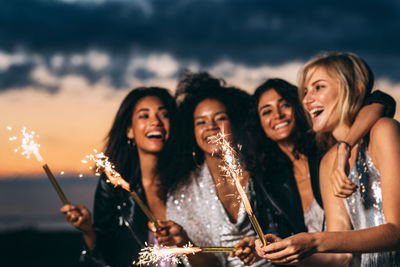 Friends holding sparklers while standing against sky at night